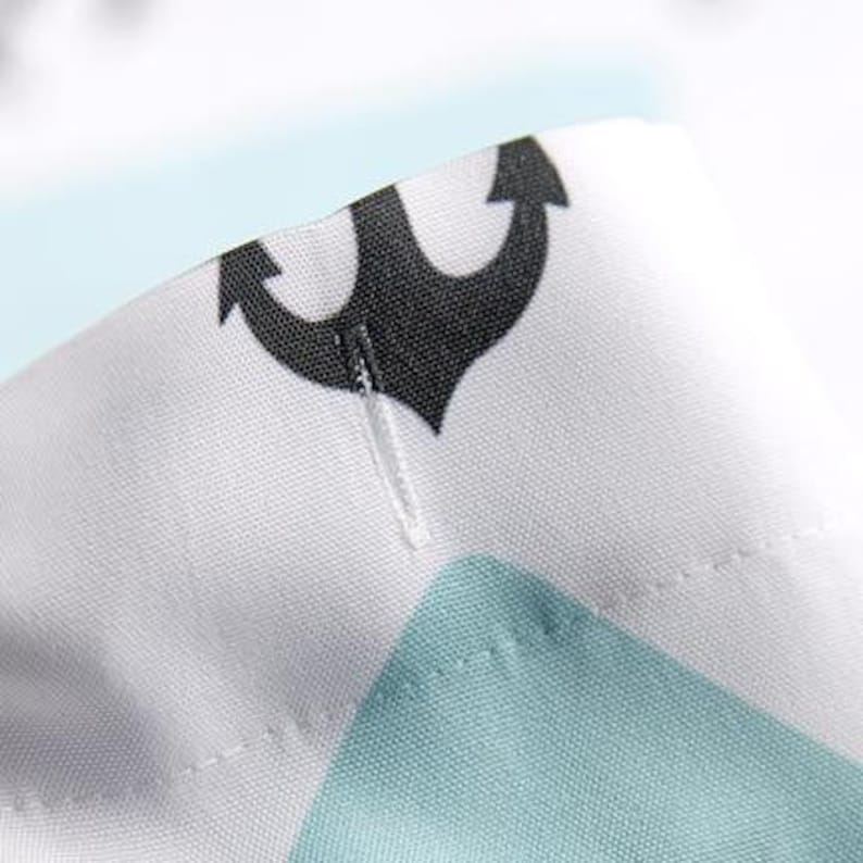 Puede incluir: Primer plano de una tela blanca con un patr&oacute;n repetido de anclas negras y formas turquesas. La tela tiene una costura visible y un gancho de pl&aacute;stico transparente. El dise&ntilde;o sugiere un tema n&aacute;utico o costero.
