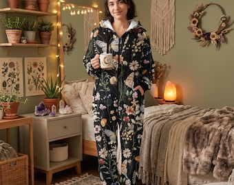 May include: A black floral and butterfly-patterned onesie with a hood and zip closure. The model is holding a mug with a crescent moon design. The background features plants, a mirror, and a bed. The onesie has pockets.
