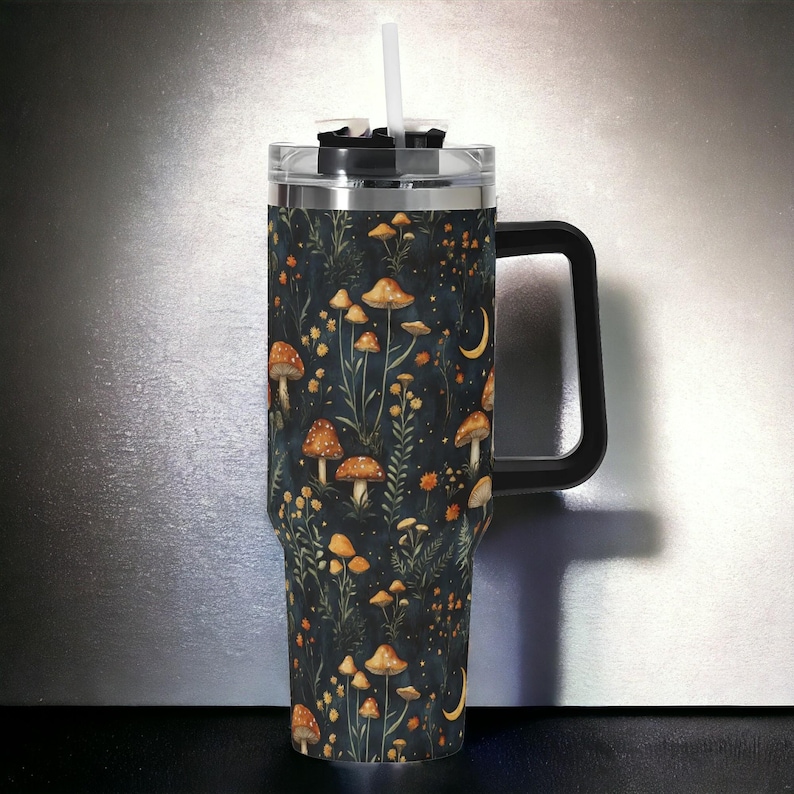 May include: A stainless steel travel mug with a black handle and lid. The mug has a dark blue background with a pattern of orange mushrooms and green leaves.