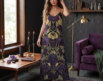 Pode incluir: Um vestido longo sem mangas com um padrão floral em tons de roxo, dourado e verde. O vestido tem alças finas e uma silhueta fluida. O fundo inclui uma mesa de madeira, velas e um sofá roxo.