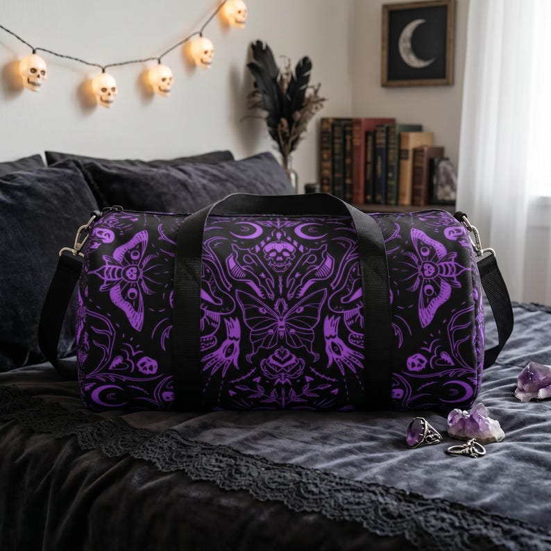May include: Black duffel bag with a purple butterfly and skull design. The bag has a black shoulder strap and handles. The design features intricate patterns and a gothic aesthetic. The bag is on a dark gray bedspread.