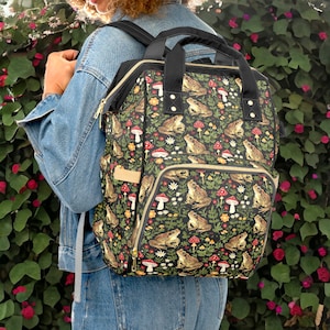 Könnte beinhalten: Ein schwarzer Rucksack mit einem Frosch- und Pilzmuster. Der Rucksack hat ein großes Hauptfach und eine kleinere Fronttasche. Der Rucksack ist aus strapazierfähigem Stoff gefertigt und hat verstellbare Träger.