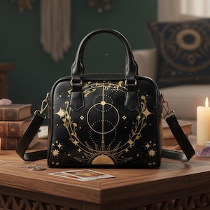 May include: Black handbag with gold celestial design, including stars, circles, and sunburst. Features a top handle and a detachable shoulder strap. The bag is sitting on a wooden surface.
