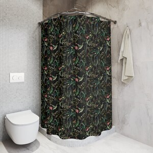 May include: Black shower curtain with a floral pattern featuring green leaves, pink and white flowers, and butterflies. The shower curtain is hanging in a bathroom with a white toilet and gray tiled walls.