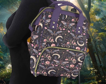 May include: A black backpack with a purple trim and a floral print featuring butterflies and moons. The print is on a dark background with pink, white, and gold accents.
