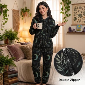 Op de afbeelding: Zwarte onesie met een hemels patroon van manen en bladeren. De onesie heeft een dubbele ritssluiting. Een persoon draagt de onesie en houdt een mok met een maanontwerp vast. De achtergrond omvat een bed, planten en decoratieve items.