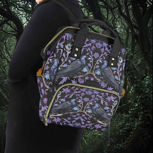 May include: A black backpack with a purple and blue floral pattern featuring black birds. The backpack has a large main compartment and a smaller front pocket.