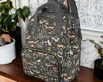 May include: A gray backpack with a black and green floral print featuring snakes, rabbits, and other woodland creatures. The backpack has a large main compartment and a smaller front pocket.