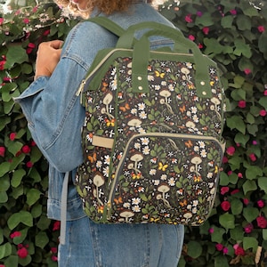 May include: A green backpack with a black and white mushroom and butterfly print. The backpack has multiple pockets and zips.