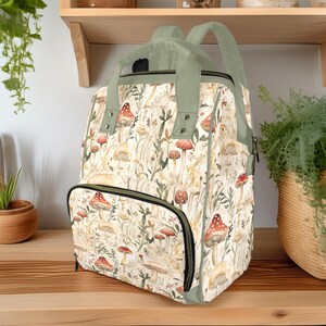 Può includere: Uno zaino beige con un motivo stravagante di funghi e fiori. La borsa è dotata di una tasca frontale con cerniera, una maniglia superiore e spallacci regolabili. Le finiture e il manico sono di colore verde tenue.