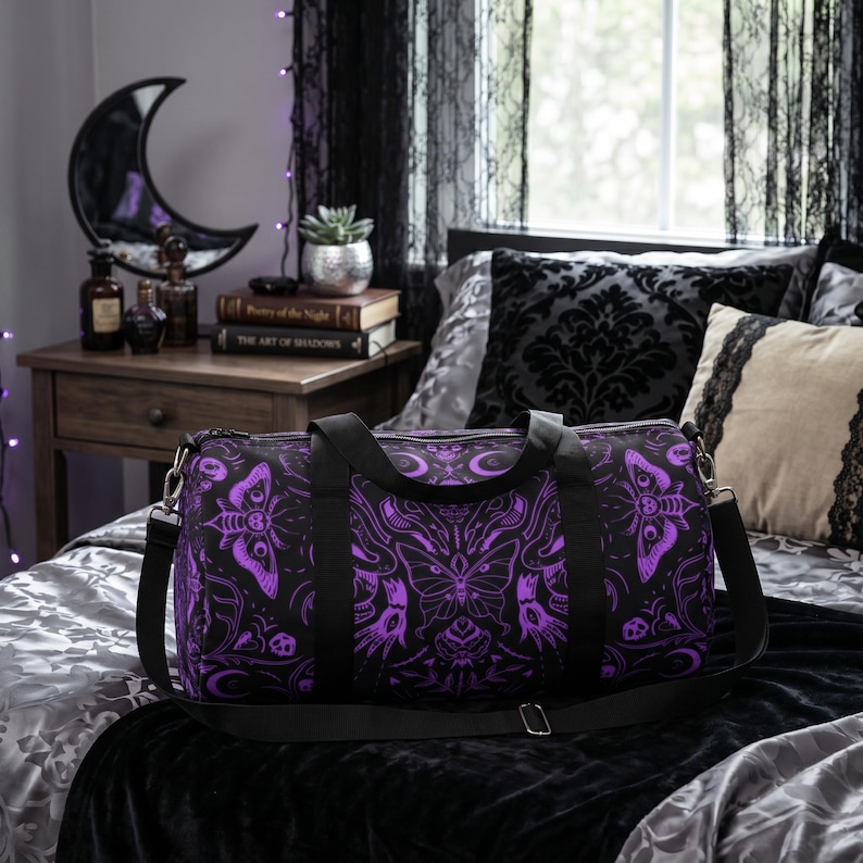 May include: Black duffel bag with a purple moth and butterfly pattern. The bag has black straps and a silver zipper. The bag is on a bed with a black velvet bedspread and decorative pillows. The room has a gothic theme.