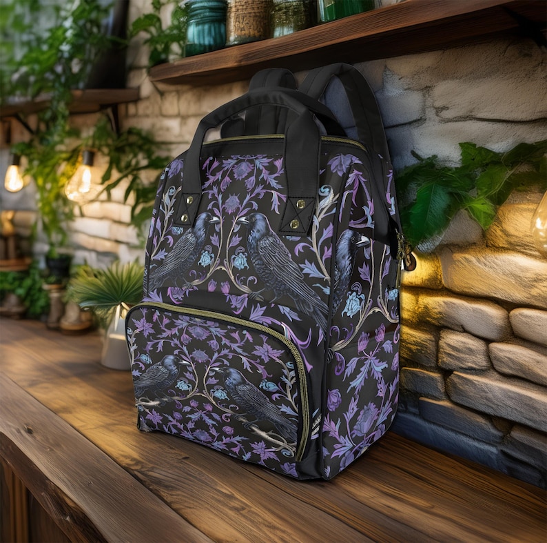 May include: A black backpack with a floral pattern featuring purple and blue flowers and black birds. The backpack has a front pocket with a zipper.