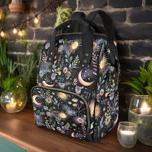 Può includere: Uno zaino nero con una stampa floreale che presenta lune, stelle e soli in tonalità dorate e viola. Lo zaino ha una grande tasca frontale con una cerniera dorata.