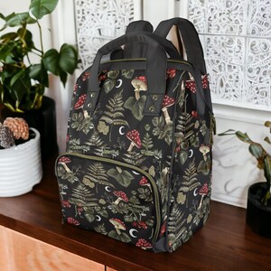 Puede incluir: Una mochila negra con un patr&oacute;n de hongos y helechos verdes y rojos. La mochila tiene un compartimento principal grande y un bolsillo frontal m&aacute;s peque&ntilde;o.