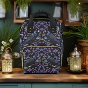 May include: A black backpack with a purple and gold floral pattern featuring black birds. The backpack has a top handle and adjustable straps.