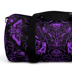 May include: Black duffel bag with a repeating purple pattern of moths, skulls, and floral designs. The bag has a black shoulder strap and a silver-colored metal zipper pull.