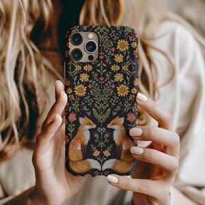 May include: A black phone case featuring a floral design with yellow sunflowers, orange and pink flowers, and two orange foxes. The case is held by a person with long blonde hair.