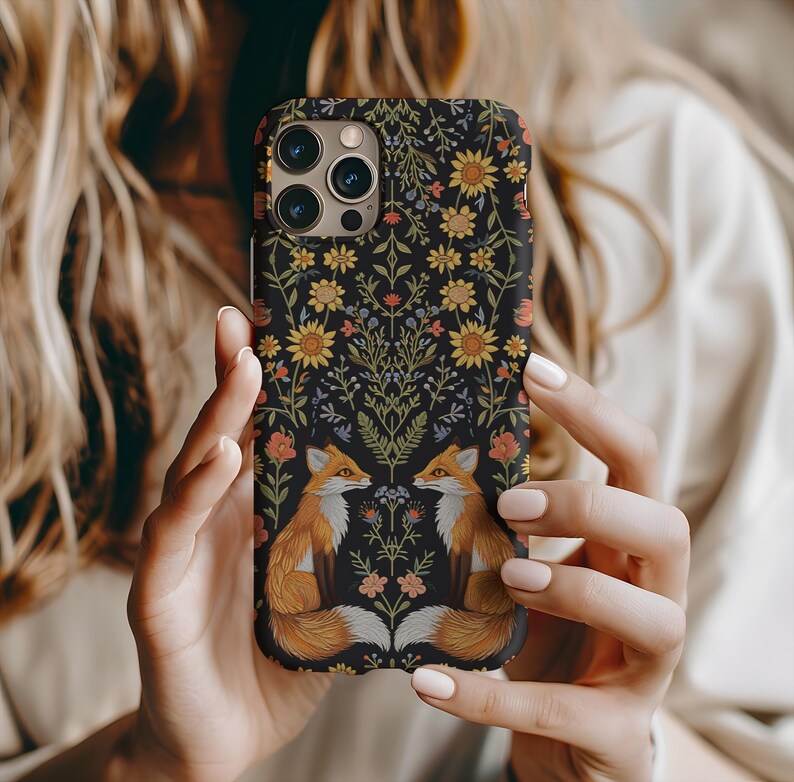 May include: A black phone case featuring a floral design with yellow sunflowers, orange and pink flowers, and two orange foxes. The case is held by a person with long blonde hair.