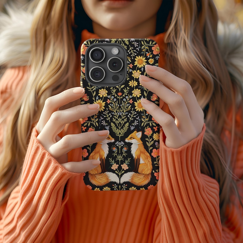 May include: A black phone case featuring a floral design with two orange foxes facing each other. The case has yellow and orange flowers and green leaves. The phone case is held by a person wearing an orange sweater.