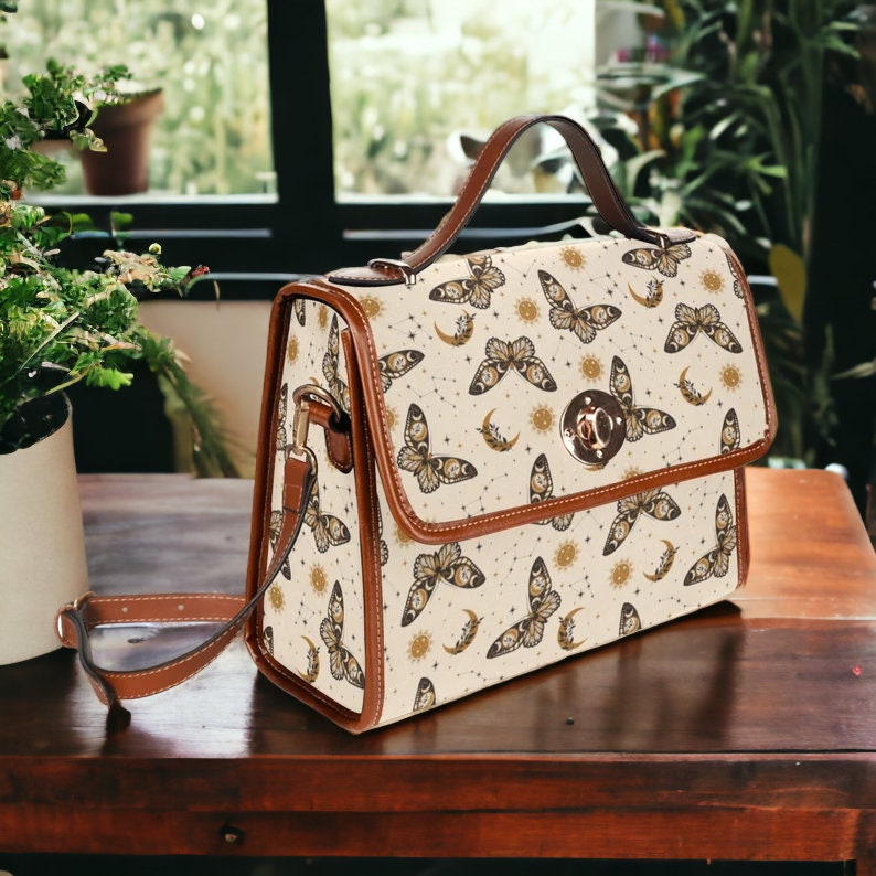 May include: A beige satchel bag with a brown leather strap and a gold clasp. The bag is decorated with a pattern of butterflies, moons, and suns.