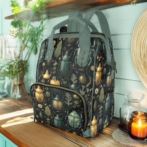 May include: A black backpack with a pattern of gold and green beetles and foliage. The backpack has gray straps and trim, a front zipper pocket, and a top handle. The bag is sitting on a wooden surface.