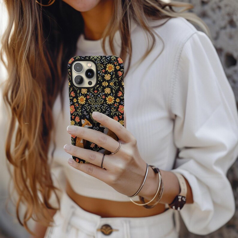 May include: Black phone case with a floral and fox design. The case features a pattern of yellow sunflowers, orange and pink flowers, and green foliage on a black background. The fox is depicted in shades of brown and white.