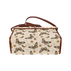 May include: A brown leather satchel bag with a beige canvas body featuring a repeating pattern of butterflies, moons, and suns. The bag has a long adjustable strap.