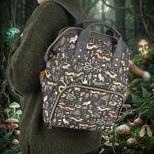 Puede incluir: Una mochila gris oscuro con un patrón repetitivo de animales del bosque, incluyendo conejos, ardillas, serpientes y caracoles, intercalados con setas, helechos y símbolos celestiales. La bolsa tiene asas superiores y un bolsillo frontal.