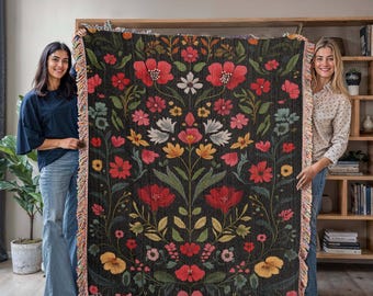 Puede incluir: Una manta de tejido de estilo tapiz con un patrón floral en tonos rojos, rosas, amarillos y verdes sobre un fondo negro. La manta tiene un borde de flecos.