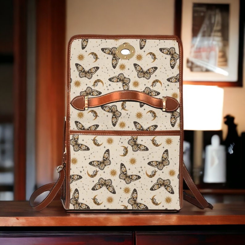 May include: A brown leather satchel with a cream canvas front featuring a repeating pattern of gold butterflies, moons, and suns. The bag has a brown leather handle and a brown leather strap.