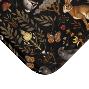Puede incluir: Una alfombra de ba&ntilde;o negra con un detallado dise&ntilde;o de bosque. El dise&ntilde;o incluye conejos marrones, mariposas, p&aacute;jaros, setas y follaje en tonos marrones, dorados y rojos. La alfombra tiene una superficie texturizada.