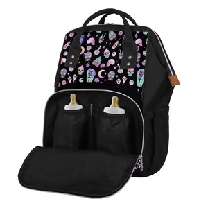 May include: Black diaper bag with a spooky and cute pattern featuring skulls, eyes, moons, and cupcakes. The bag has two bottle holders on the front and a large zippered pocket on the bottom.