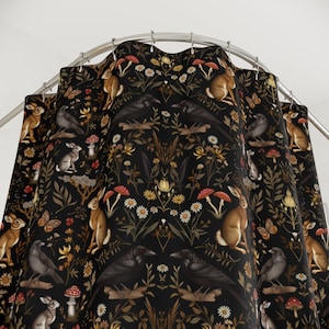 Puede incluir: Cortina de ducha negra con un patr&oacute;n repetido de conejos, cuervos, setas, mariposas y flores. El dise&ntilde;o incluye elementos marrones, rojos, amarillos y blancos sobre un fondo oscuro. La cortina cuelga de una barra met&aacute;lica curva.
