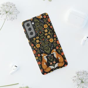 May include: Black phone case featuring a floral pattern with orange, yellow, and red flowers. Two foxes are depicted on the case, with orange fur and white accents. The case is designed for a smartphone.