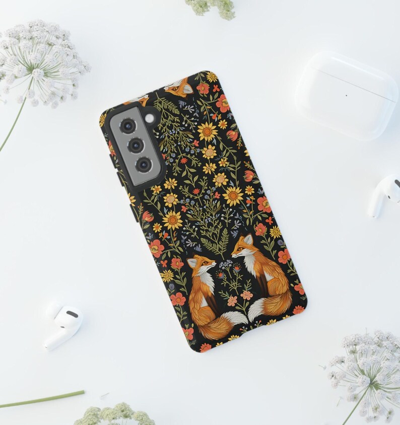 May include: Black phone case featuring a floral pattern with orange, yellow, and red flowers. Two foxes are depicted on the case, with orange fur and white accents. The case is designed for a smartphone.