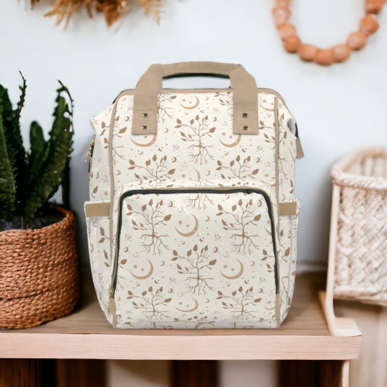 May include: A cream-colored backpack with a repeating pattern of brown trees, leaves, moons, and stars. The bag has a tan handle and trim, with a front pocket and side pockets. The bag is sitting on a wooden surface.