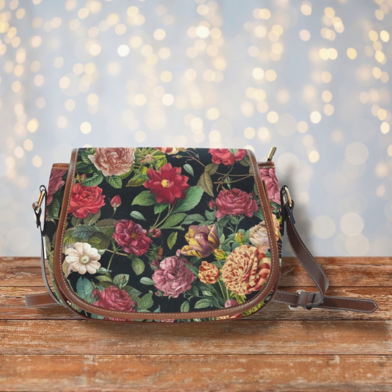 Op de afbeelding: Een bruine leren schoudertas met een bloemenprint met roze, rode en witte bloemen op een zwarte achtergrond. De tas heeft een lange verstelbare schouderband.
