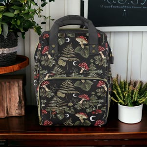 Puede incluir: Una mochila negra con estampado de setas verdes y rojas. La mochila tiene un compartimento principal grande y un bolsillo frontal m&aacute;s peque&ntilde;o. La mochila est&aacute; hecha de un tejido resistente y tiene correas ajustables.