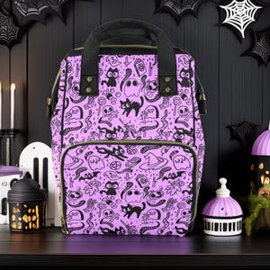 May include: A purple backpack with a black and white Halloween-themed print. The print features cats, ghosts, skulls, bats, and other spooky imagery. The backpack has a black zipper and a black handle.