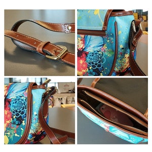 Op de afbeelding: Een blauwe en bruine canvas schoudertas met een bloemen- en verfspatten ontwerp. De tas heeft een bruine leren band en een ritssluiting. De tas is open om de binnenvoering en een ritsvak te laten zien.