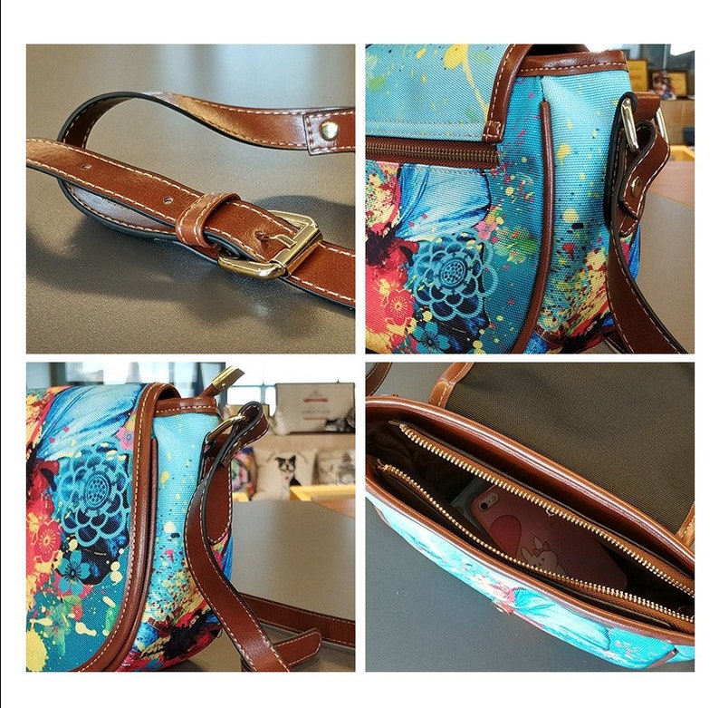 Op de afbeelding: Een blauwe en bruine canvas schoudertas met een bloemen- en verfspatten ontwerp. De tas heeft een bruine leren band en een ritssluiting. De tas is open om de binnenvoering en een ritsvak te laten zien.
