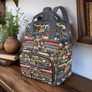 Può includere: Uno zaino grigio con stampa floreale e di libri. Lo zaino ha una tasca frontale con cerniera, spallacci grigi e una maniglia superiore. Il motivo floreale comprende fiori rosa, gialli e bianchi. Lo sfondo della stampa è una pila di libri.