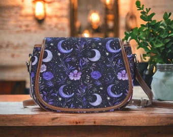 May include: A purple and brown satchel bag with a crescent moon and floral pattern. The bag has a long adjustable strap.
