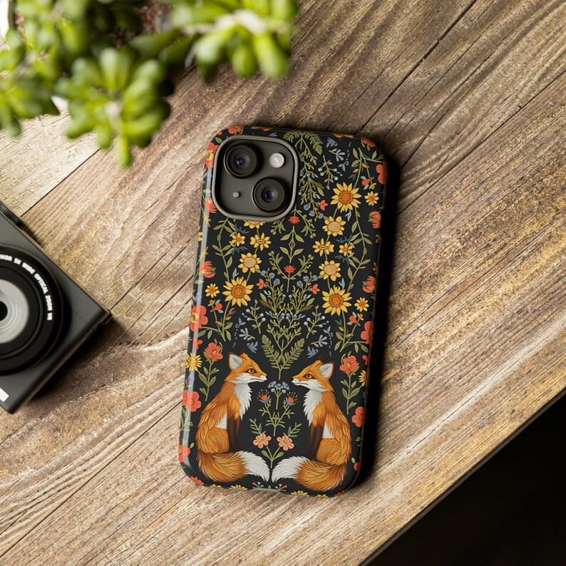 May include: A black phone case featuring a floral design with two foxes facing each other. The case has a glossy finish and is decorated with yellow, orange, and blue flowers. The phone case is on a wooden surface.