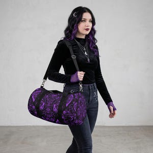 May include: A black and purple duffel bag with a detailed, gothic-inspired pattern. The bag has a cylindrical shape with a black shoulder strap and handles. The design features intricate illustrations in a dark purple hue against a black background, creating a striking contrast.
