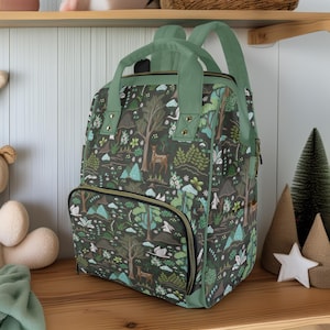 Puede incluir: Una mochila verde oscuro con un estampado de bosque con árboles, ciervos y montañas. La mochila tiene correas verdes, un asa superior y un bolsillo frontal con cremallera. La bolsa está sobre una estantería de madera.