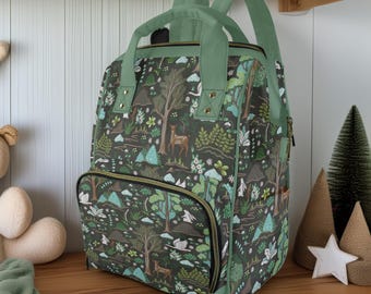 May include: A dark green backpack with a forest pattern featuring trees, deer, and mountains. The backpack has green straps, a top handle, and a front pocket with a zipper. The bag is sitting on a wooden shelf.
