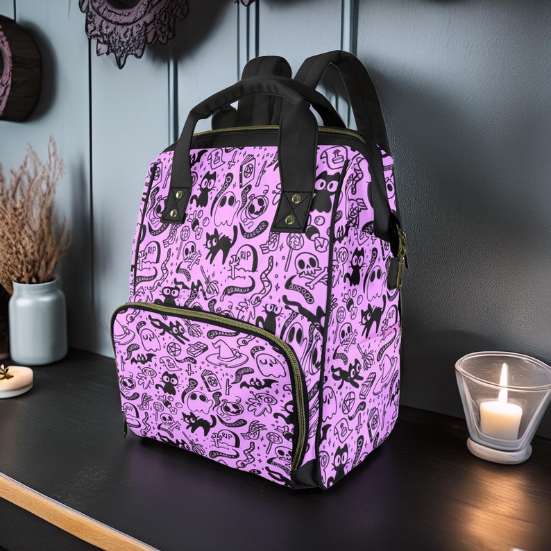 May include: A purple backpack with a black and white print of ghosts, cats, and other Halloween-themed imagery. The backpack has a large main compartment and a smaller front pocket.