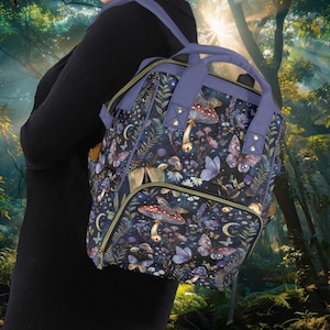 Può includere: Uno zaino viola e nero con una scena di foresta fantasiosa con funghi, farfalle e fasi lunari. Lo zaino ha una tasca con cerniera e spalline regolabili.