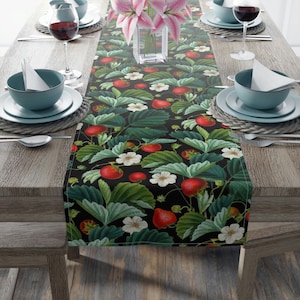 May include: A black table runner with a repeating pattern of red strawberries, green leaves, and white flowers. The runner is on a wooden table with two place settings on each side.